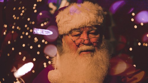 A close-up of Santa Claus with a fluffy white beard, glasses, and a festive background of colorful lights.