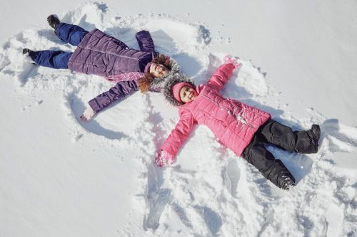 Two children in colorful winter coats making snow angels in a snowy landscape.