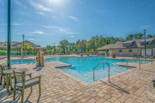 A sunny outdoor pool area with lounge chairs, surrounded by trees and buildings, featuring clear blue water and a bright sky.