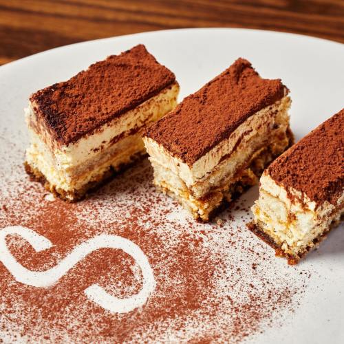 Three rectangular slices of tiramisu dusted with cocoa powder on a white plate, with a decorative swirl of cocoa.