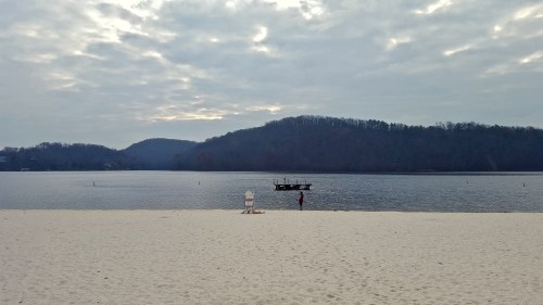 A serene beach scene with a calm lake, a distant dock, and a person standing on the shore under a cloudy sky.