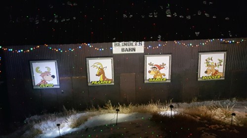 A barn decorated with colorful lights featuring cartoon reindeer: Cupid, Prancer, Dasher, and Donner. Snow on the ground.