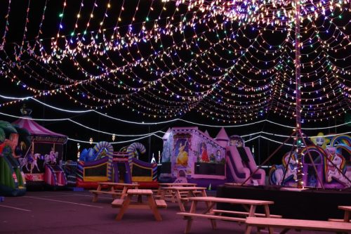 Colorful lights illuminate a festive outdoor area filled with inflatable attractions and picnic tables.