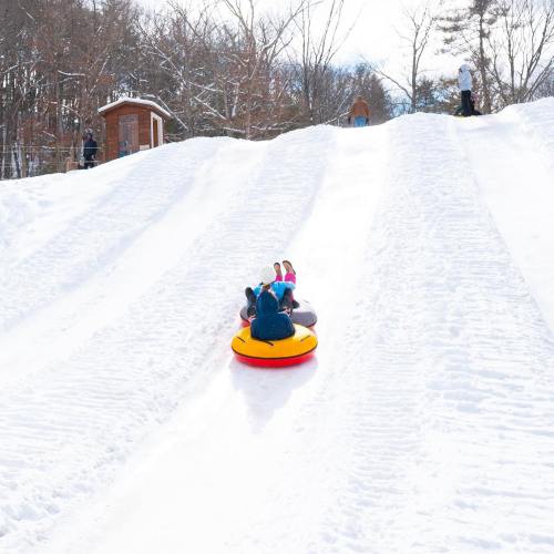 Two people slide down a snowy hill on inflatable tubes, with trees and a small building in the background.