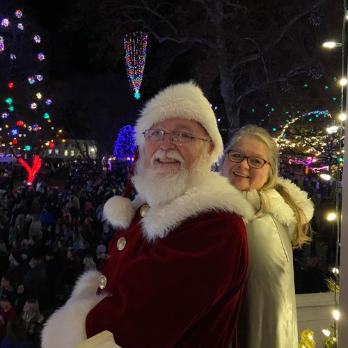 A smiling Santa Claus in a festive setting with colorful lights, standing next to a woman with glasses, surrounded by a crowd.