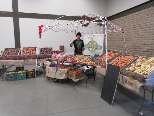 A festive market stall with various vegetables and a person in an elf costume standing under a decorated canopy.