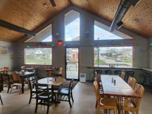 Interior of a cozy restaurant with wooden ceilings, tables, and large windows showing a street view outside.