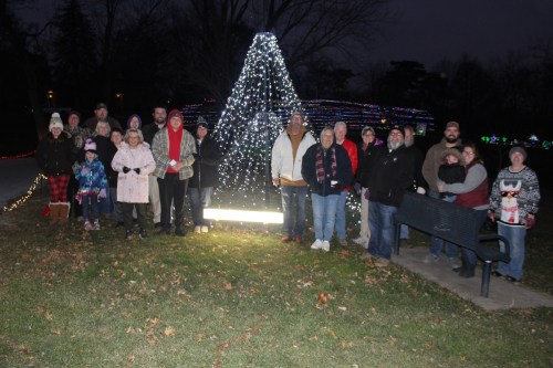 A group of people stands together in front of a lit Christmas tree, surrounded by festive holiday lights at dusk.