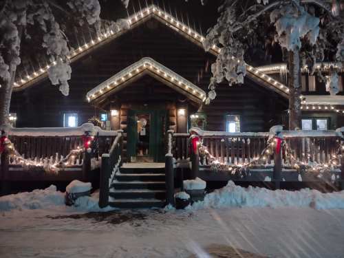 A cozy log cabin decorated with holiday lights and snow, featuring a welcoming entrance and festive garlands.