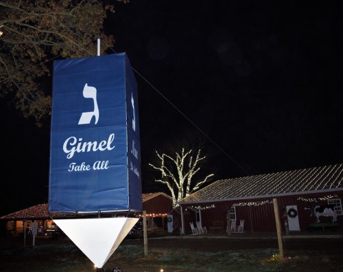 A large blue banner with a white dreidel symbol and the word "Gimel" hangs outside a building decorated with lights.