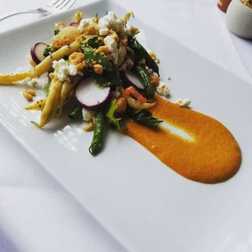 A colorful salad with green beans, radishes, and crumbled cheese, drizzled with a vibrant orange sauce on a white plate.