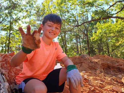 A smiling boy in an orange shirt shows a dirty hand while sitting on a rocky surface in a wooded area.