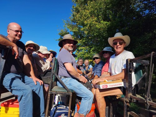 A group of people sitting together outdoors, wearing hats and smiling, enjoying a sunny day.