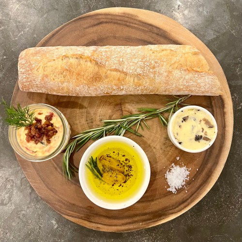 A wooden platter with a baguette, olive oil, flavored butter, and a dip, garnished with herbs and salt.
