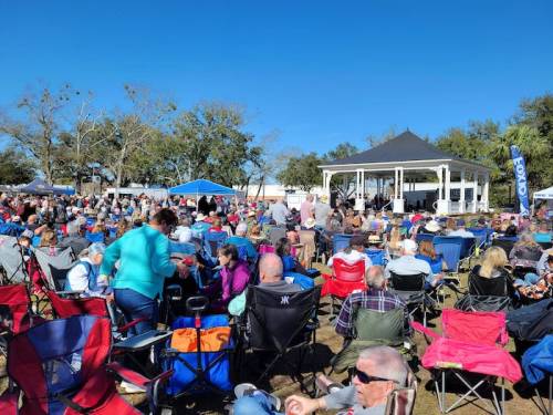 A large crowd gathers in a park for an outdoor event, with many people seated in folding chairs under a clear blue sky.