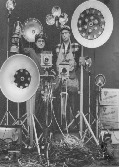 Two photographers stand among vintage lighting equipment and cameras, surrounded by cables and studio gear.