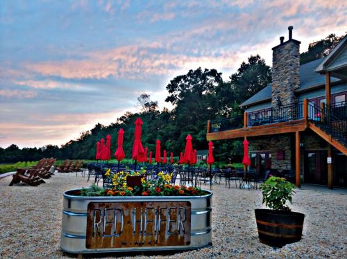 A rustic outdoor dining area with red umbrellas, flower planters, and a stone building under a colorful sky at sunset.