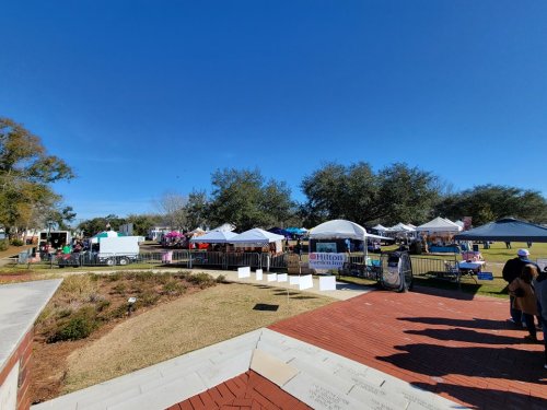 A vibrant outdoor market with tents and vendors under a clear blue sky, surrounded by trees and grassy areas.