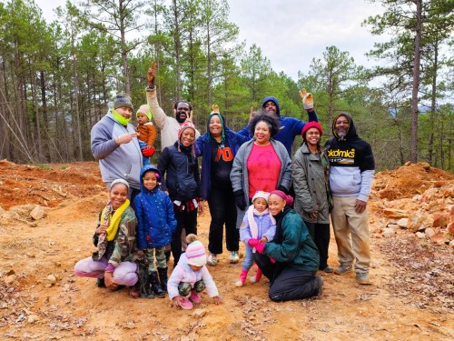 A diverse group of people, including children and adults, smiling and posing together outdoors in a forested area.