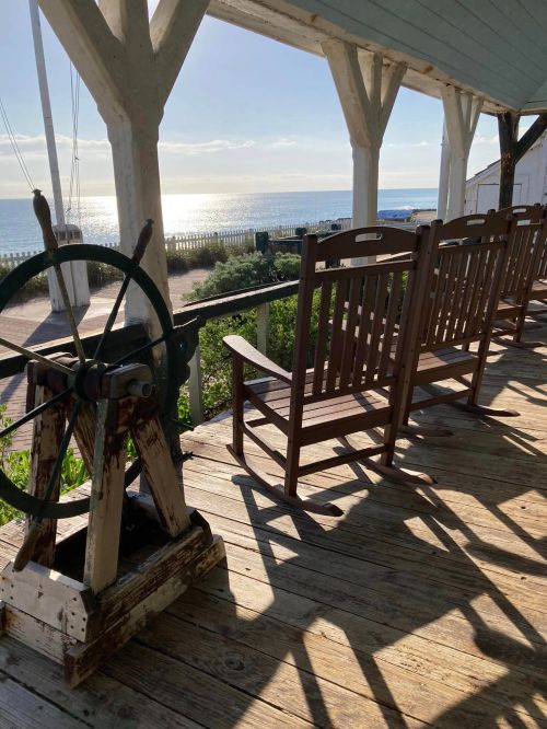 A wooden porch with rocking chairs overlooking a calm sea and bright sky, featuring a ship's wheel in the foreground.