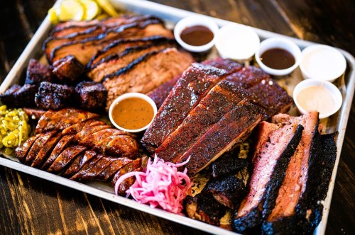 A platter of assorted smoked meats, including brisket, sausage, and ribs, served with various sauces and pickled onions.