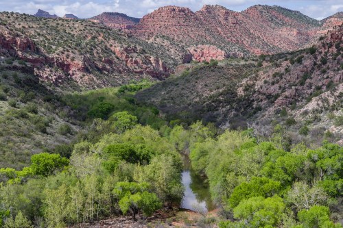 A lush green valley with a winding river, surrounded by red rock mountains and hills under a partly cloudy sky.
