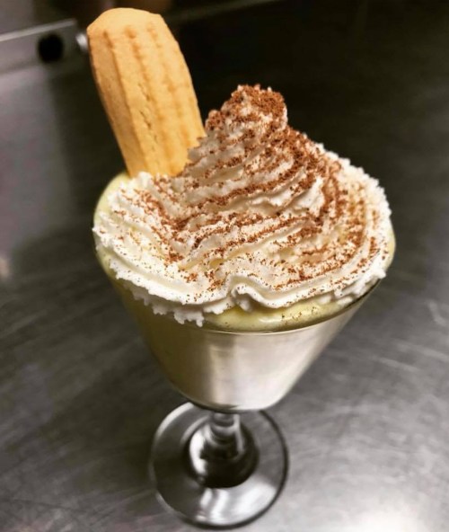 A dessert in a glass topped with whipped cream, cocoa powder, and a cookie, set on a stainless steel surface.
