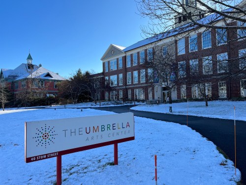 A brick building with a sign reading "The Umbrella Arts Center" in a snowy landscape.