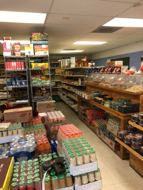 A well-stocked grocery store aisle with shelves of snacks, drinks, and canned goods, plus boxes on the floor.