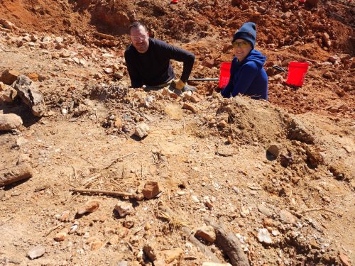 Two people working at a dig site, surrounded by dirt and rocks, with red buckets in the background.