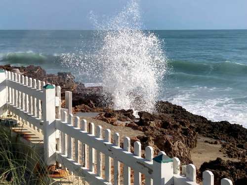 Waves crashing against rocky shore near a white picket fence, with a clear blue sky in the background.