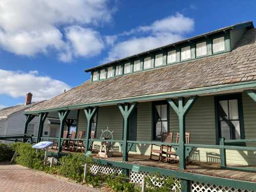 A green wooden house with a porch, rocking chairs, and a blue sky with clouds in the background.