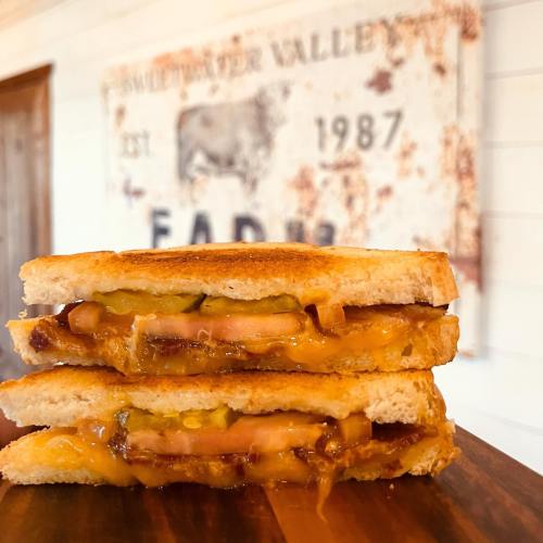A stacked sandwich with melted cheese, pickles, and layers of meat on toasted bread, set against a rustic background.