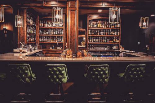 A stylish bar with wooden shelves filled with bottles, green upholstered stools, and warm lighting.