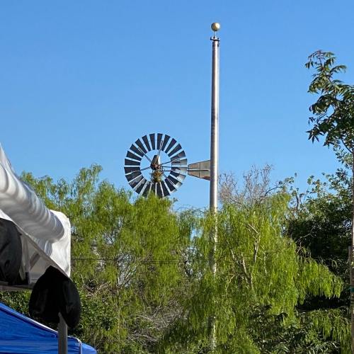 A windmill stands tall against a clear blue sky, surrounded by green trees and a flagpole nearby.