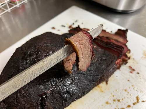 A close-up of a slice of smoked brisket on a knife, resting on a cutting board with the whole brisket in the background.