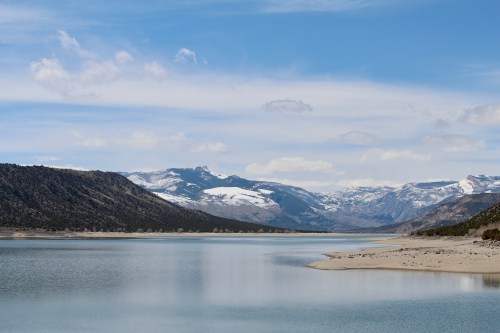 A serene lake surrounded by mountains, with snow-capped peaks under a blue sky and fluffy clouds.