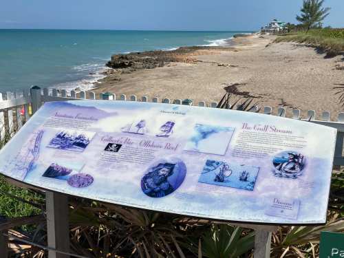 Informational sign by the beach, detailing local history and marine life, with a view of the ocean and shoreline.