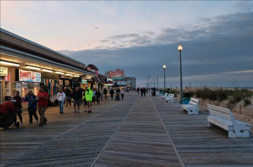 A bustling boardwalk with shops and people walking, set against a cloudy sky at dusk.