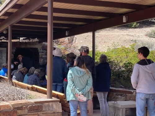 A group of people gathered under a shelter, listening to a speaker in a natural setting.