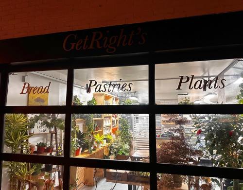 A storefront window displays the words "Bread," "Pastries," and "Plants" with a cozy interior filled with greenery.