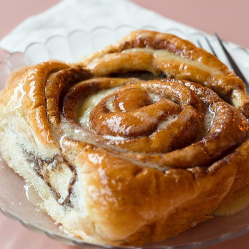A close-up of a freshly baked cinnamon roll drizzled with icing, served on a glass plate.