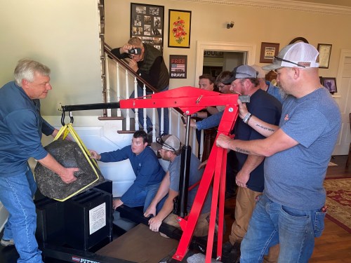 A group of people using a hoist to move a large stone inside a house, with stairs and framed pictures in the background.