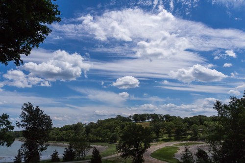 A scenic view of a lake surrounded by trees under a bright blue sky with fluffy white clouds.