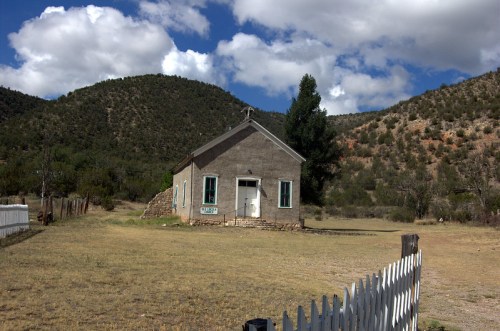 A small, weathered house with green windows sits in a grassy field, surrounded by mountains and a white picket fence.