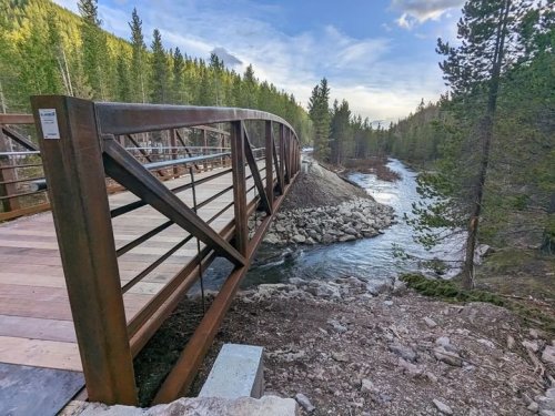 A metal arch bridge spans a river, surrounded by trees and rocky terrain under a clear sky.