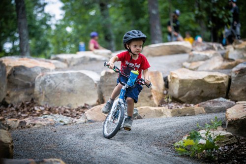 A young child in a helmet rides a bike on a rocky trail surrounded by trees and other cyclists.