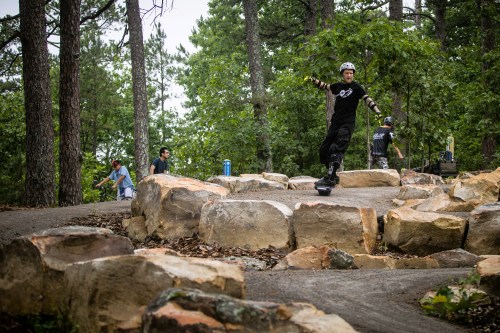 A person skateboarding on a winding path surrounded by trees and rocks, with others in the background.