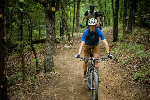 Two mountain bikers navigate a dirt trail through a lush, green forest. One rider is in the foreground.