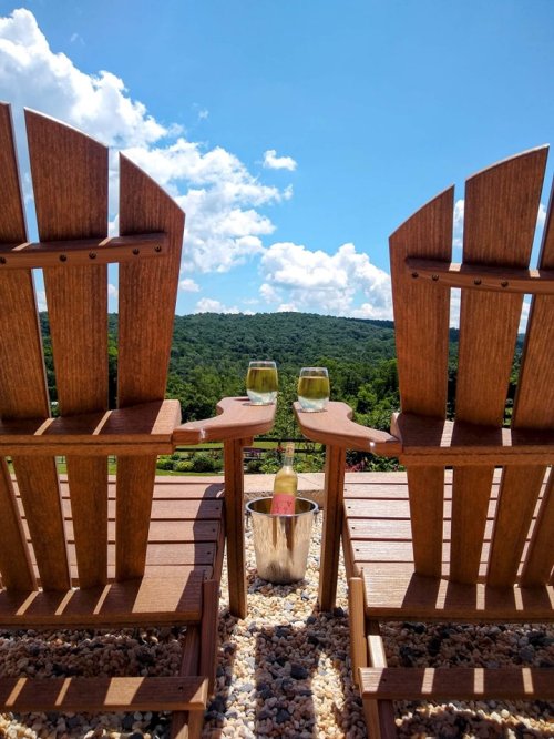 Two wooden chairs facing a scenic view, with two glasses of wine and a champagne bucket between them under a blue sky.